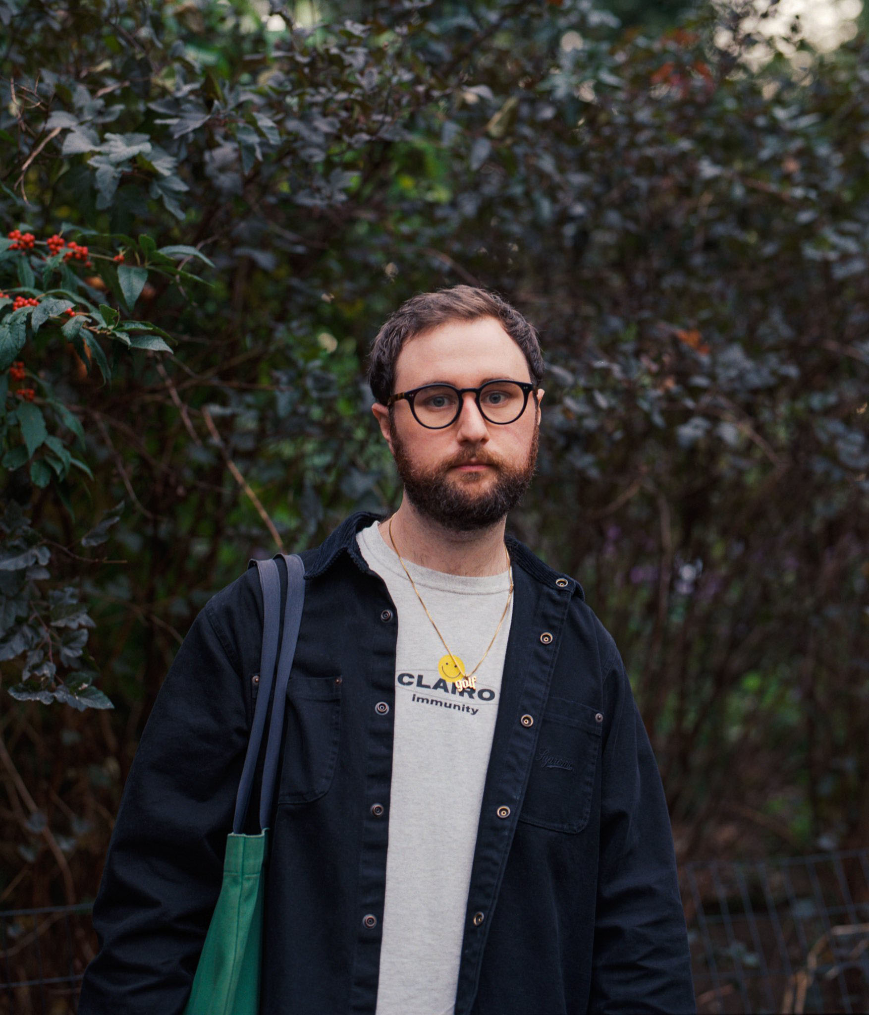 Ronin wearing a Clairo t-shirt and black jacket with a green tote bag in front of bushes.